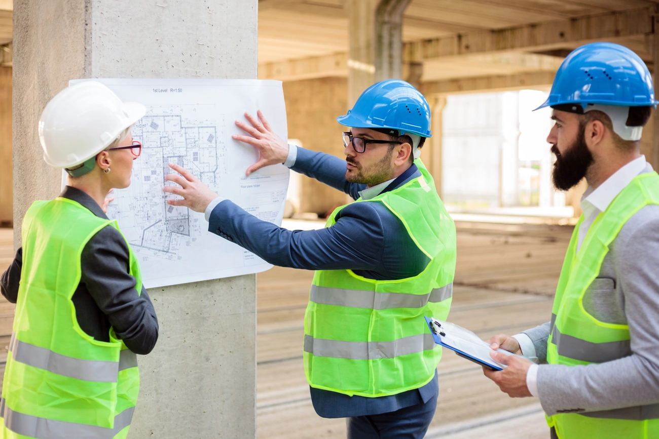 M-J tief.und Rohrleitungsbau GmbH - Team erfolgreicher junger Architekten und Geschäftspartner während eines Meetings auf einer Baustelle. Betrachten von Architekturzeichnungen und Bauplänen.